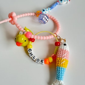 May include: A pink beaded bracelet with a yellow rubber duck, a blue dice charm, and a crocheted fish charm. The bracelet also has white letter beads spelling out the name "CHLOE".