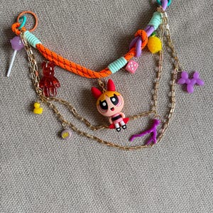 Puede incluir: Un collar colorido con una variedad de dijes, que incluyen un personaje de dibujos animados, un osito de goma, un dado, un limón, un caniche y una piruleta. El collar está hecho de cuerda naranja, cadena dorada y cuerda morada.