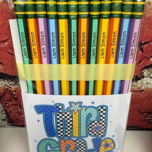 May include: A set of twelve multi-colored pencils with green metal ferrules and pink erasers, each personalized with the name Liam Simon in black text. The pencils are held together by a yellow paper band inside a clear plastic sleeve with a card featuring the text Third Grade Squad in a checkered and colorful graphic font.