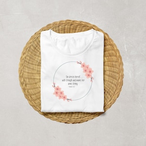 以下が含まれることがあります： ピンクの花柄がデザインされた白いTシャツ。"彼女は強さで身を飾り、腕を強くする。箴言31:21"というテキストが書かれている