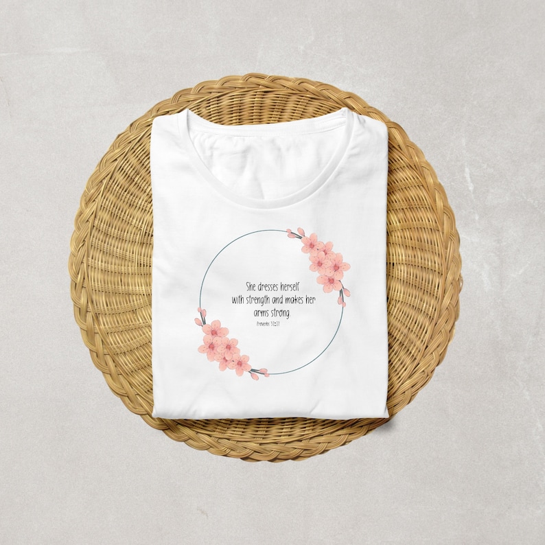 以下が含まれることがあります： ピンクの花柄がデザインされた白いTシャツ。"彼女は強さで身を飾り、腕を強くする。箴言31:21"というテキストが書かれている