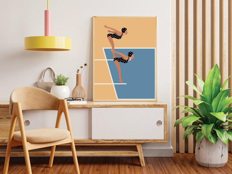 May include: A framed illustration of two women in black and white polka dot swimsuits diving off a diving board into a blue pool. The background is a light orange color.