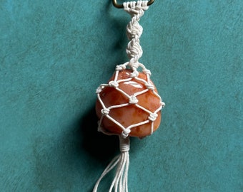 May include: A white macrame keychain with a peach-colored stone in the center. The keychain has a gold metal clasp.