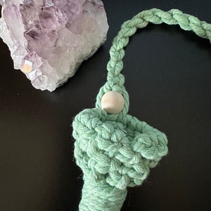 May include: A light green macrame tassel with a wooden bead at the top. The tassel is hanging from a light green cord.
