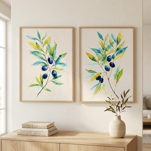 May include: Two framed watercolour prints of olive branches with green and yellow leaves and dark blue olives. The prints are in light brown wooden frames. A small vase with olive branches sits on a wooden cabinet below the prints.