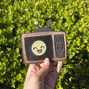 May include: A patch in the shape of a vintage television set. The TV has a black screen with a smiling yellow face. The border is a metallic gold color, and the antenna is black. The patch is held against a background of green foliage.