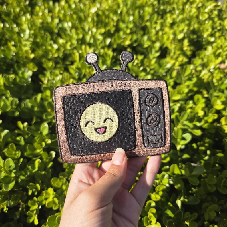 May include: A patch in the shape of a vintage television set. The TV has a black screen with a smiling yellow face. The border is a metallic gold color, and the antenna is black. The patch is held against a background of green foliage.