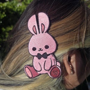 May include: A pink embroidered bunny patch with a black bow tie. The bunny has large, round eyes and is sitting down. The patch is attached to a person's hair. The patch is a cute accessory.