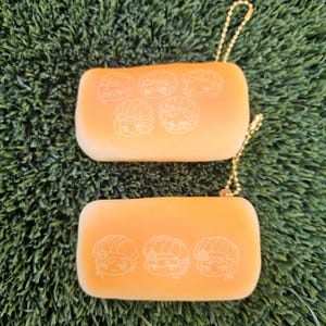 May include: Two rectangular orange pouches with gold chain pulls. Each pouch has a series of white cartoon character faces printed on the front. The pouches are lying on a bed of green artificial grass.