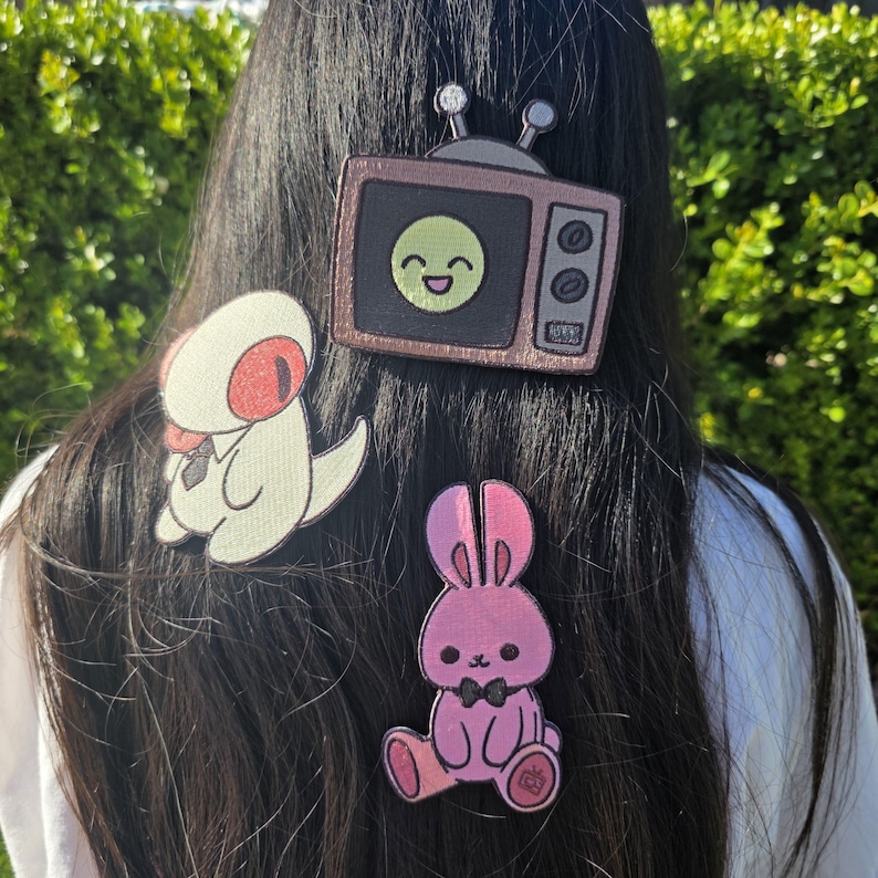 May include: Three embroidered patches are attached to dark hair. A retro TV with a smiling face, a white dinosaur with a red eye, and a pink bunny with a black bow tie. The patches are colorful and whimsical.
