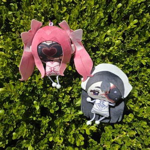 May include: Two plush character charms featuring stylized designs. One charm has pink hair with bows and a heart-shaped face mask. The other charm has black hair, a white head covering, and a face mask with a red mark. Both have white string limbs.