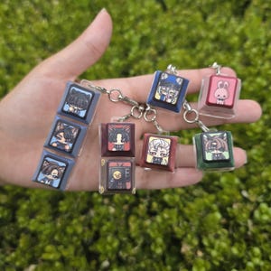 May include: A collection of keycap keychains featuring various anime-style character illustrations. The keycaps are square, colored in shades of blue, maroon, red, and green, and attached to silver metal rings. Each keycap displays a unique graphic.