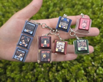 Puede incluir: Una colección de llaveros con forma de teclas de teclado que presentan diversas ilustraciones de personajes estilo anime. Las teclas son cuadradas, de colores azul, granate, rojo y verde, y están unidas a anillas de metal plateado.