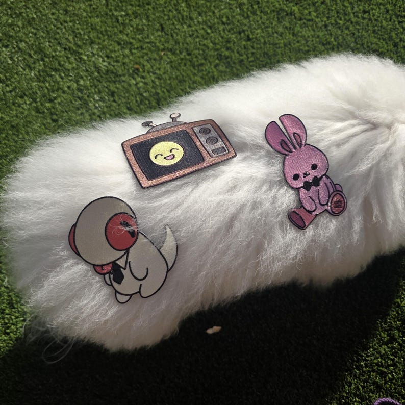May include: Three embroidered patches: a retro television with a smiling face, a pink bunny wearing a bow tie, and a cartoon character in a spacesuit. The patches are on a white, fluffy surface, set against a green background.