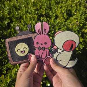 May include: Three embroidered patches featuring cartoon characters. One is a brown television with a smiling face, another is a pink bunny with a bow tie, and the third is a light green creature with a red eye.