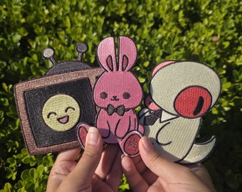 Puede incluir: Tres parches bordados con personajes de dibujos animados. Uno es un televisor marrón con una cara sonriente, otro es un conejito rosa con pajarita y el tercero es una criatura verde claro con un ojo rojo.
