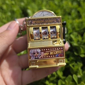 May include: A gold-colored slot machine keychain featuring three spinning reels with illustrated character faces. The base displays a cartoon figure with white hair and the text "EARN 800 COINS FOR VIP ACCESS" alongside red and blue poker chip graphics.