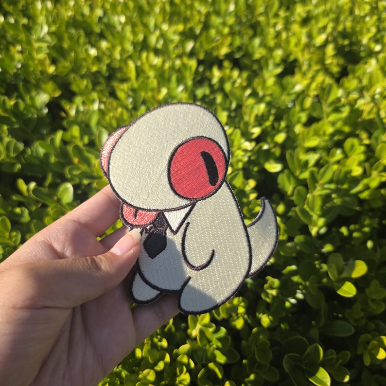 May include: Embroidered patch featuring a cartoon creature with a white body, red eyes, and a black tie. The patch has a stitched border and is held against a backdrop of green foliage. The design is whimsical and playful.
