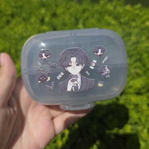 May include: A translucent, oval-shaped plastic storage container featuring a central sticker of an anime-style character in a suit, surrounded by smaller stickers of various chibi figures, candies, and a small creature with antlers.
