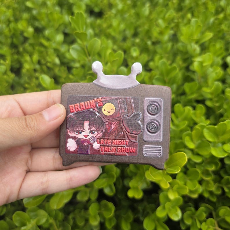 Puede incluir: Un pin marr&oacute;n con forma de televisor de dibujos animados con una antena blanca. El pin presenta un gr&aacute;fico de una persona sosteniendo un micr&oacute;fono con el texto "Braun's Late Night Talk Show". El pin tiene una cara sonriente en la pantalla del televisor.