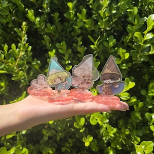 May include: Four acrylic character stands featuring cartoon wizards with different colored hats and robes. Each stand is on a pink circular base. The background is a lush green bush.