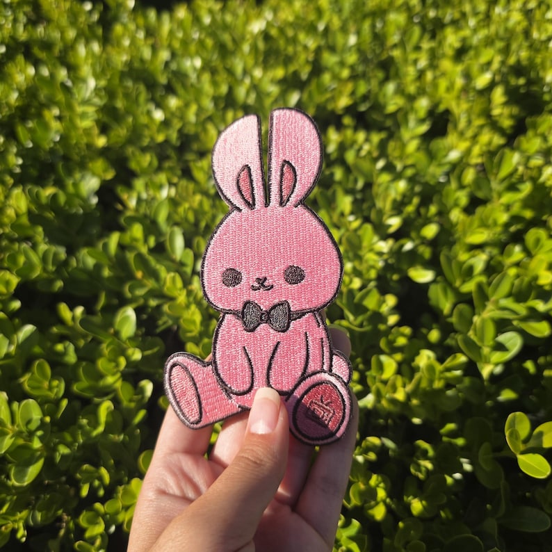 May include: A pink bunny patch with a black bow tie and embroidered details. The patch features a sitting bunny design with a red emblem on its foot. The patch is held in front of a green leafy background.