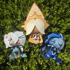 Könnte beinhalten: Drei Plüschfiguren mit stilisierten Designs. Eine Figur hat weißes Haar und einen grünen Umhang, eine andere trägt einen hohen gelben spitzen Hut und helles Haar, und die dritte hat blaues Haar und ein dunkelblaues Outfit mit schwarzen Akzenten.