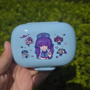 May include: A light blue rectangular plastic storage case featuring a central illustration of a purple-haired character in a nurse-style outfit, surrounded by four smaller chibi-style character graphics and scattered pill-shaped icons.