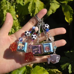 Puede incluir: Una colección de llaveros hechos a mano con ilustraciones de personajes de estilo anime en miniatura dentro de cubos de resina transparente. Cada llavero tiene una cuenta de balón de fútbol, una rosa y un dije de tiburón. Los llaveros están unidos a un llavero plateado.