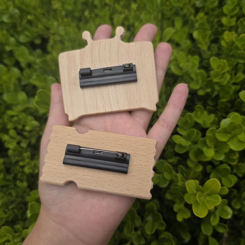Puede incluir: Dos placas de identificaci&oacute;n de madera con respaldos de alfiler negros. Una placa tiene una parte superior redondeada con dos peque&ntilde;os recortes decorativos, la otra es rectangular con un borde festoneado. Las placas son de color marr&oacute;n claro.