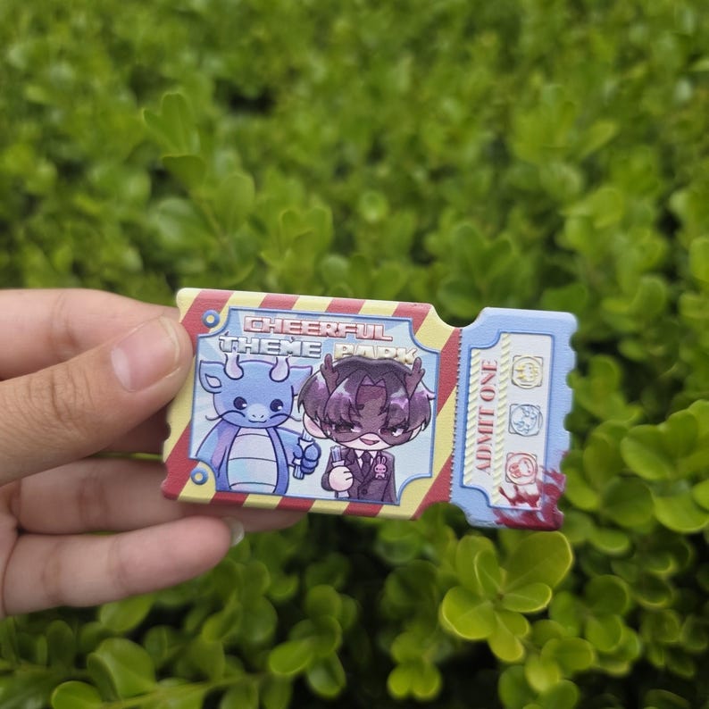 Puede incluir: Pin rectangular con forma de boleto con el texto "Cheerful Theme Park" y "Admit One". El pin presenta ilustraciones de dibujos animados de dos personajes, un drag&oacute;n azul y una persona con traje. El pin tiene un esquema de color rojo, amarillo y azul.