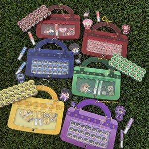 May include: A collection of colorful, handbag-shaped display cases with clear windows, each filled with small character badges. The cases are in various colors, including red, blue, green, yellow, and purple, and are arranged on a green surface.