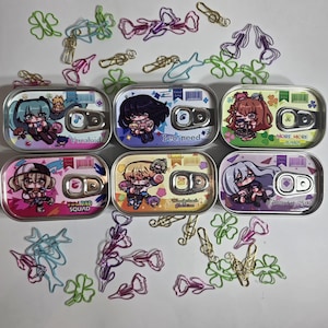 May include: Six decorative tin cans with anime-style character illustrations and text. The cans feature colorful designs and the text "Vocaloid", "Leo/need", "MORE MORE JUMP", "Vivid BAD SQUAD", and "ntcard at 25:00". Various decorative paperclips are scattered around the cans.