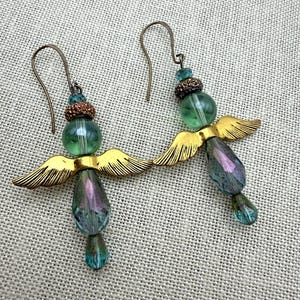 May include: A pair of dangle earrings featuring a green glass bead, a small bronze-toned textured bead, and a teardrop-shaped iridescent blue-green crystal bead. Each earring includes a gold-toned metal wing component and a curved ear wire hook.