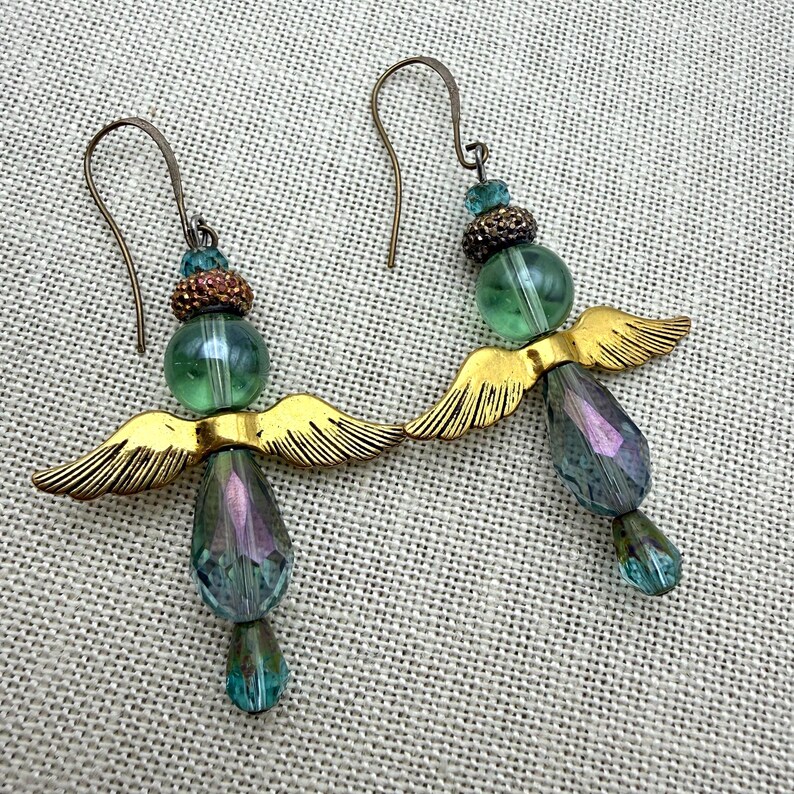 May include: A pair of dangle earrings featuring a green glass bead, a small bronze-toned textured bead, and a teardrop-shaped iridescent blue-green crystal bead. Each earring includes a gold-toned metal wing component and a curved ear wire hook.