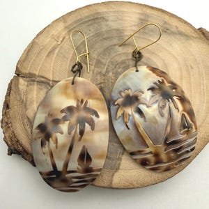 May include: Oval-shaped earrings with a tropical beach scene carved into the surface. The earrings feature palm trees and a sailboat, with a warm color palette of browns and tans. The ear wires are gold-colored.