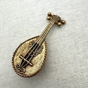 May include: A gold-toned metal brooch shaped like a lute or mandolin musical instrument. The piece features a textured, pear-shaped body with a rope-style border, a long neck with tuning pegs at the top, and raised string and bridge details.