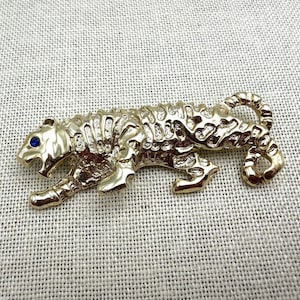 May include: A gold-toned metal brooch shaped like a prowling tiger with a textured, striped body and a single small, round blue stone set into the eye. The tiger is depicted in a walking stance with its tail curled upward and back toward its body.