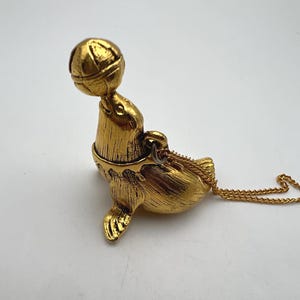 May include: A gold-colored pendant necklace featuring a seal balancing a ball on its nose. The seal is detailed with textured fur and flippers. The necklace has a delicate gold chain.