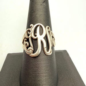 May include: A silver-toned metal ring featuring a large, ornate, cut-out monogram design. The centerpiece consists of stylized, interconnected cursive letters that form a decorative pattern with swirling flourishes and smooth, polished edges.