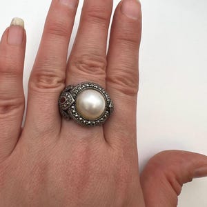 May include: A silver ring featuring a large, round, white pearl at the center. The pearl is surrounded by a border of small, dark stones. The ring has an ornate design with additional small, red accents.