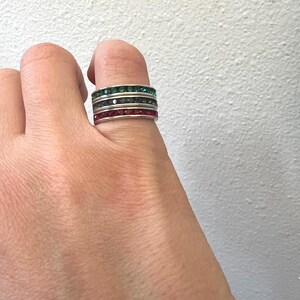 May include: A set of four silver rings with colorful gemstones. The rings are stacked on each other, with the top ring having green gemstones, the second ring having black gemstones, the third ring having clear gemstones, and the bottom ring having red gemstones.