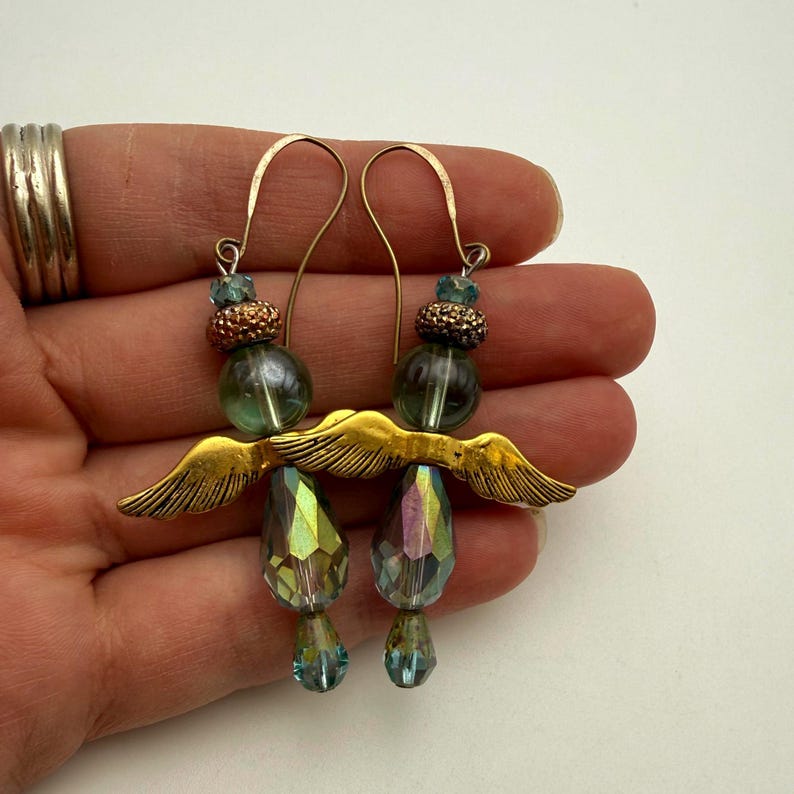 May include: A pair of dangle earrings featuring gold-toned metal wings, round translucent green beads, textured copper-coloured spacers, and faceted iridescent teardrop-shaped glass crystals suspended from curved gold-toned metal ear wires.