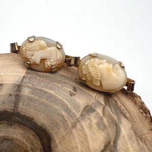 May include: A gold-toned metal brooch featuring two oval-shaped cameos. Each cameo displays a carved cream-colored profile of a person facing left, set against a translucent beige background. The two cameos are connected by a central metal link.