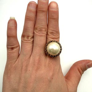 May include: A large, round, cream-colored faux pearl ring with a gold-tone beaded setting. The ring is shown on a hand, highlighting its size and design. The ring's setting has a textured, detailed appearance.