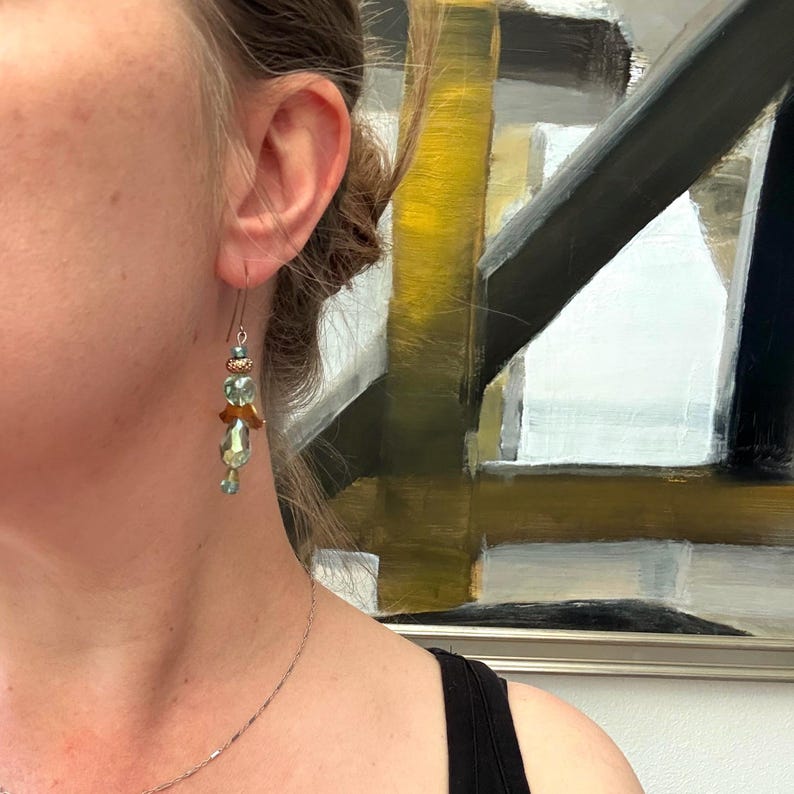 May include: A dangle earring featuring a thin metal hook, a small metallic bead, a light green faceted glass bead, a copper-coloured wing-shaped component, and a larger light green teardrop-shaped glass bead hanging at the bottom of the earring.