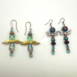 May include: Two pairs of drop earrings. The first pair features gold-tone wings, light green beads, and iridescent teardrop crystals. The second pair features silver-tone wings, dark purple and teal beads, and small blue crystal accents.
