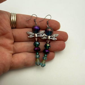 May include: A pair of dangle earrings featuring silver-toned dragonfly charms. Each earring is composed of a large iridescent purple bead at the top, followed by smaller faceted beads in shades of green, purple, and blue, ending in a teardrop bead.