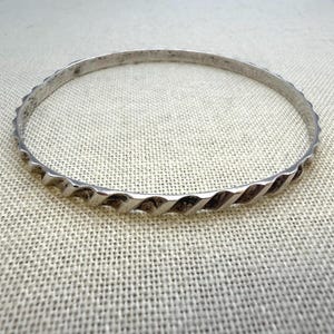 May include: A circular silver bangle bracelet featuring a continuous twisted rope pattern along its entire outer circumference. The metal surface has a polished finish with subtle darkened areas within the grooves of the twisted design.
