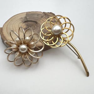 May include: Two gold-toned flower brooches with pearl accents. One brooch features a layered petal design, while the other has a circular pattern. The brooches are displayed on a wooden surface.